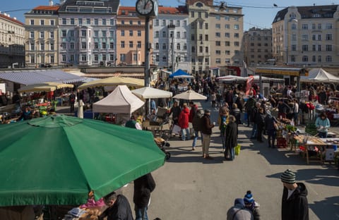 Mercado de Karmelitermarkt num sábado, em Viena, distrito de Leopoldstadt, com bancas, vendedores, transeuntes e edifícios urbanos no fundo.