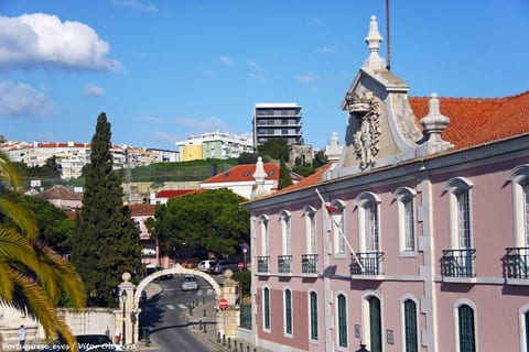 Núcleo antigo de Oeiras, com edifício histórico cor-de-rosa e arco em pedra em primeiro plano e, na colina ao fundo, blocos de habitação coletiva.