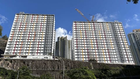 Duas torres de 18 pisos de habitação pública em Hong Kong com fachadas de módulos empilhados e uma grua no topo, semanas antes da conclusão da obra.